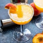 Peach margarita in stemmed glass rimmed with salt. A bottle of tequila is in the background.
