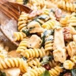 Wooden spoon lifting creamy sun dried tomato chicken pasta out of a skillet.