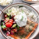 White bowl filled with rice and topped with salmon, tzatziki, a Greek salad, and feta.