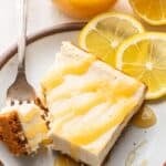 Lemon cheesecake slice drizzled with lemon curd on a white plate with a piece cut off and laying on a fork.
