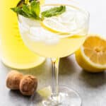 A limoncello spritz in a coupe glass with a bottle of limoncello and a halved lemon are behind it.