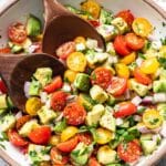 Tomato avocado salad in a white bowl with wooden serving spoons.