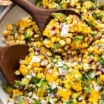 Avocado Corn Salad in a white bowl with wooden serving spoons scooping it up.