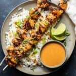 Gray plate with Two Thai chicken skewers on top of rice and a bowl of peanut sauce next to them.