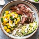 Sliced steak, mango cucumber salsa, and avocado on top of rice in a white bowl.
