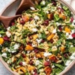 Butternut squash orzo salad with spinach, dried cranberries, goat cheese and pepitas in a white serving bowl with wooden spoons.