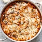 Parmesan chicken and orzo topped with melted cheese in a skillet. A small bowl of parmesan cheese is behind the skillet.
