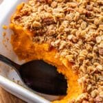 Sweet potato casserole with streusel topping in a rectangular white casserole dish. A scoop is missing and a black serving spoon is in the dish.