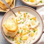 Chicken pot pie soup in a white bowl with a spoon and a biscuit on top of the soup. A pan of biscuits is behind the bowl of soup.