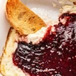 Baked goat cheese dip topped with raspberry chipotle sauce and a crostini in the baking dish with some of the dip on it.