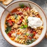 Italian sausage pasta soup in a white bowl with a spoon in it and a dollop of ricotta on top.