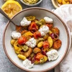 Pesto gnocchi with sausage, peppers, and tomatoes in a white bowl topped with ricotta.