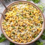 Mexican street corn salad in a white serving bowl with spoons. Cotija cheese and limes next to it.