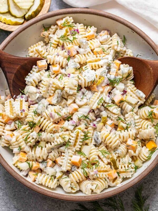 Dill pickle pasta salad in a white serving bowl with two wooden serving spoons in the bowl.