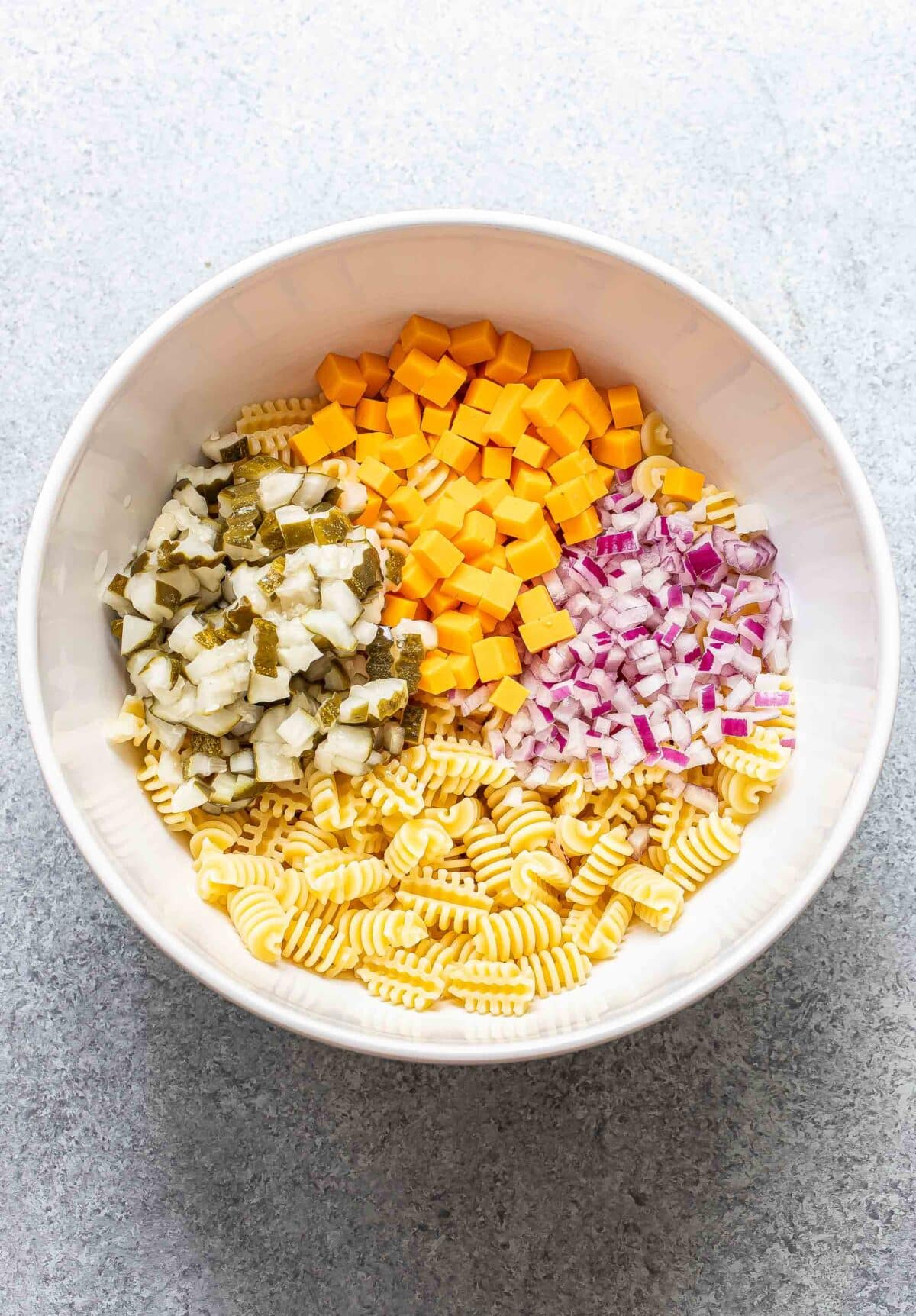 Pasta, cheddar cheese, pickles, and red onion in a white mixing bowl.