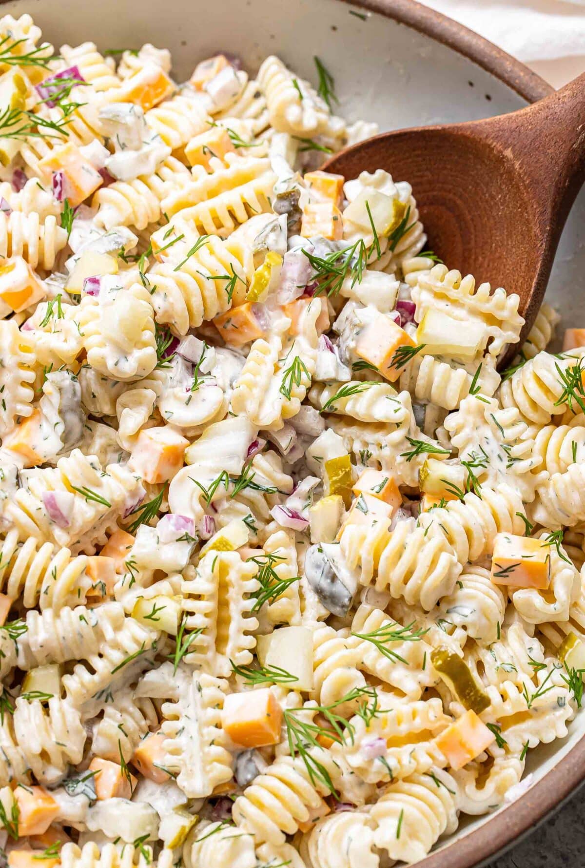 Pickle pasta salad in a bowl with a wooden spoon scooping it up.
