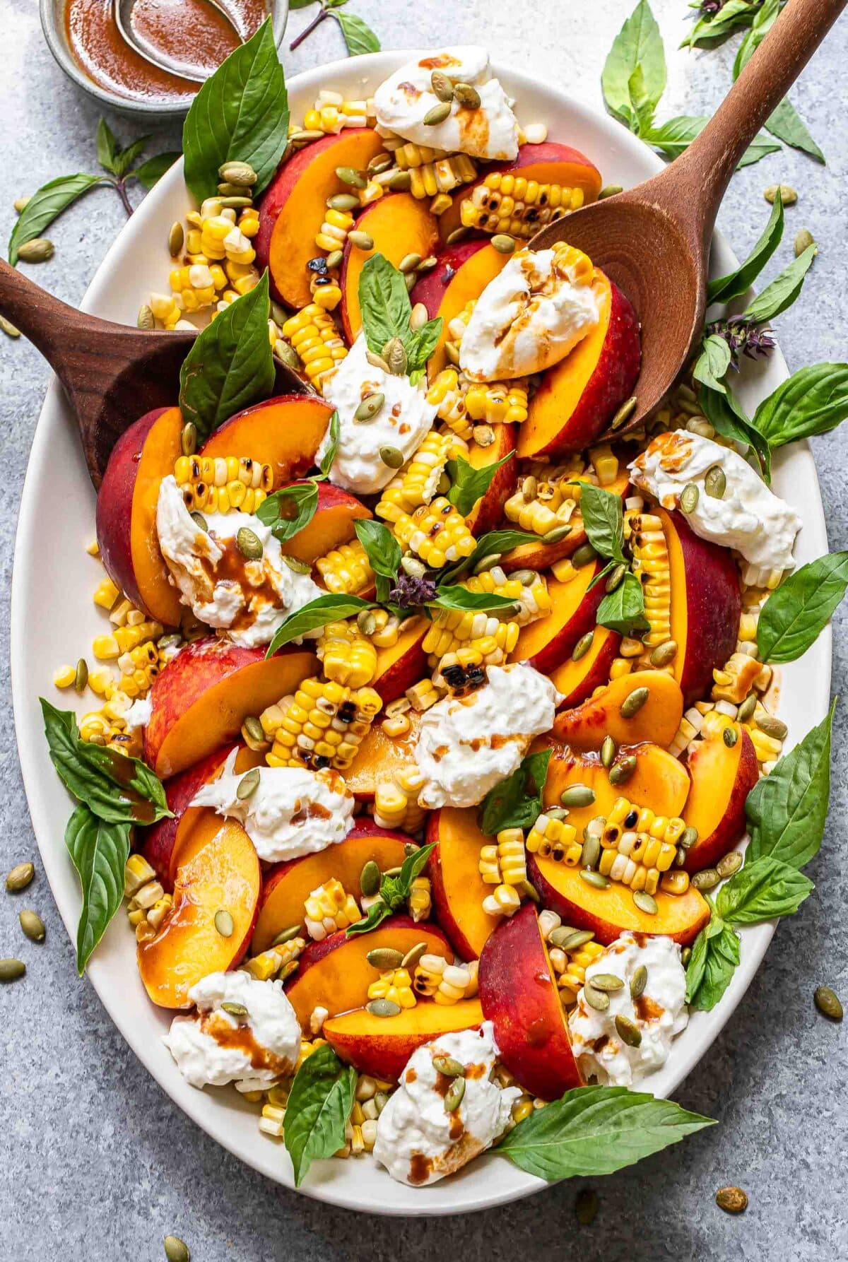 Burrata peach salad with corn and basil on an oval serving plate with wooden serving spoons.