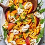 Burrata peach salad with corn and basil on an oval serving plate with wooden serving spoons.