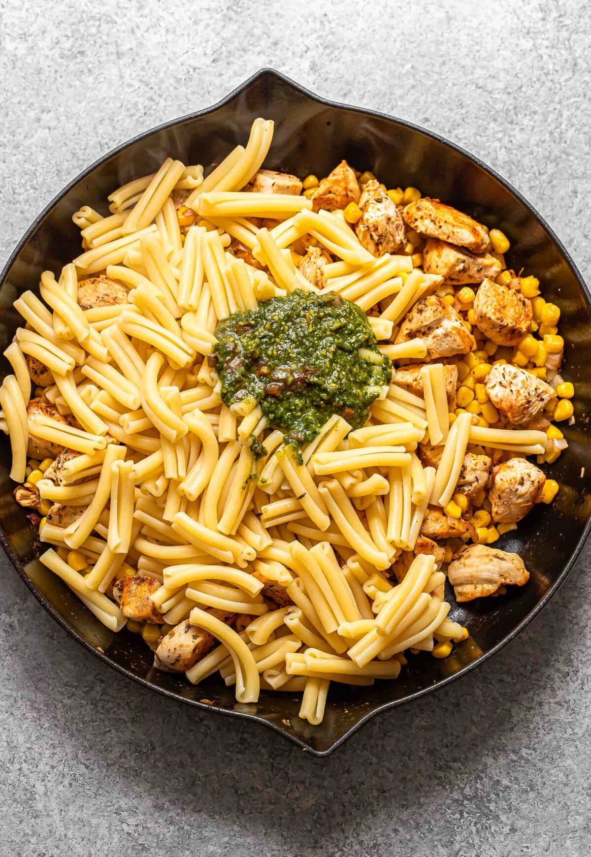 Chicken, corn, pasta and pesto in a skillet before being stirred together.
