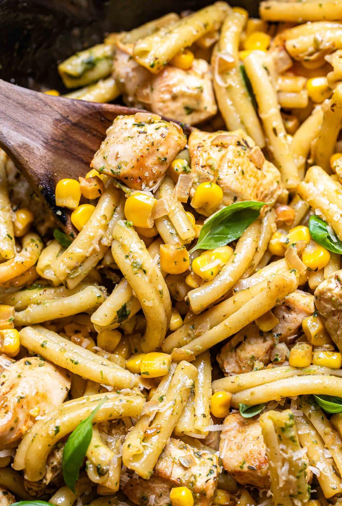 Chicken and corn pasta being lifted out of a skillet with a wooden serving spoon.