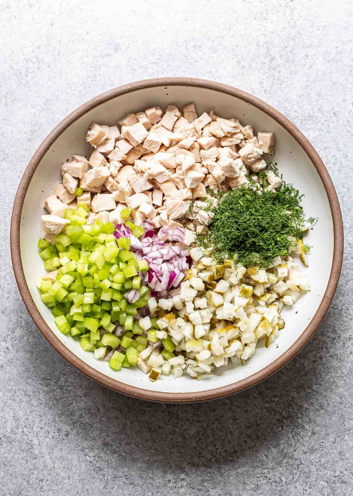 Chicken, celery, red onion, pickles, and dill in a white bowl.