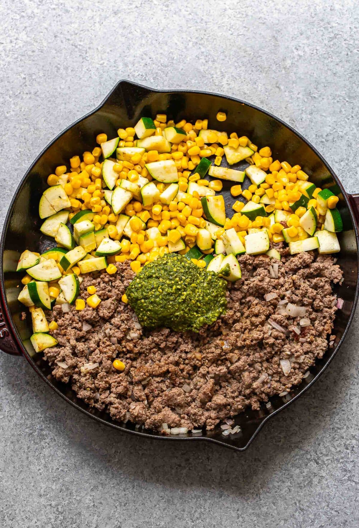 Ground beef, zucchini and corn in a skillet with a dollop of pesto on top.