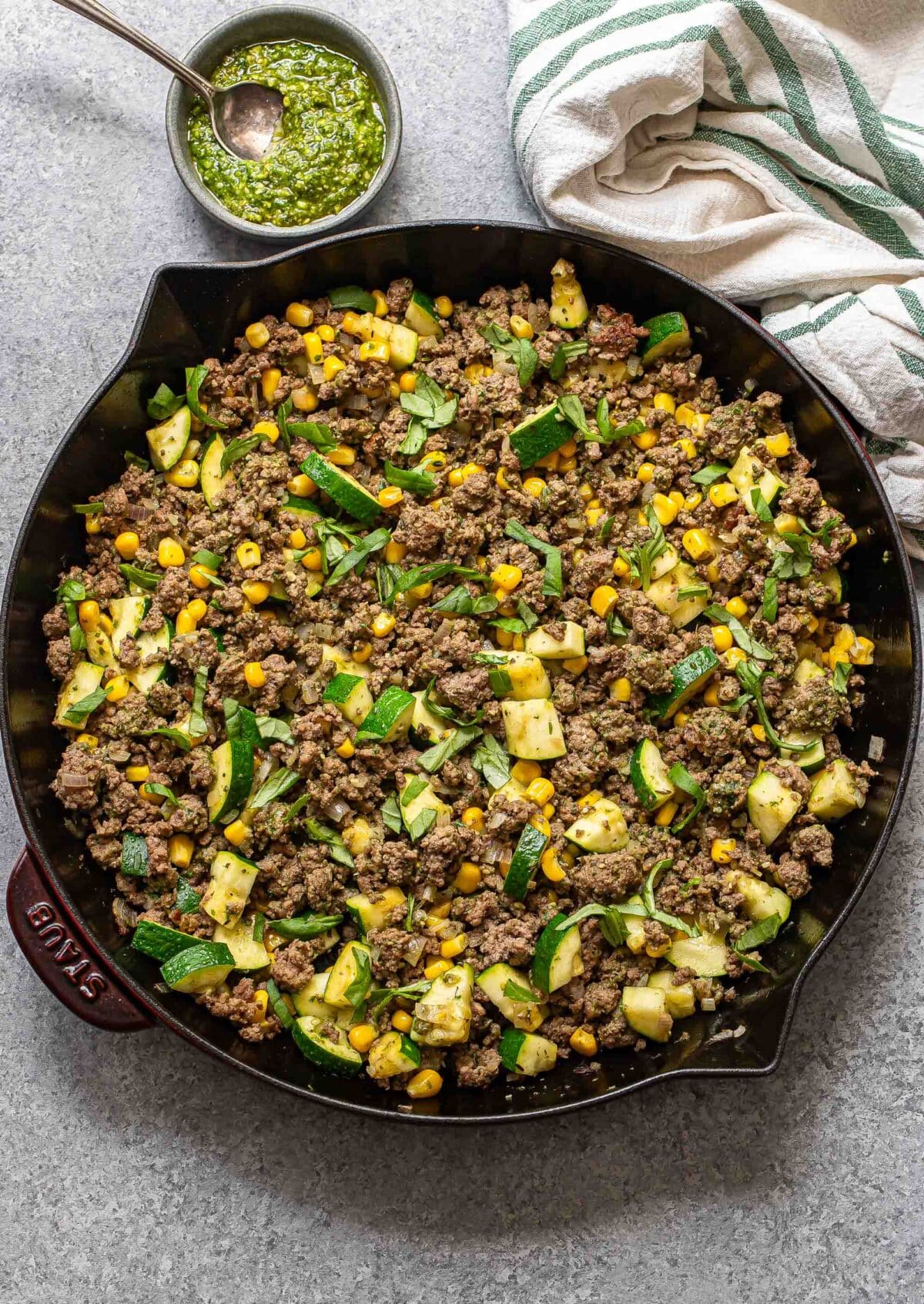 Pesto ground beef with zucchini and corn in a skillet with a bowl of pesto behind it.