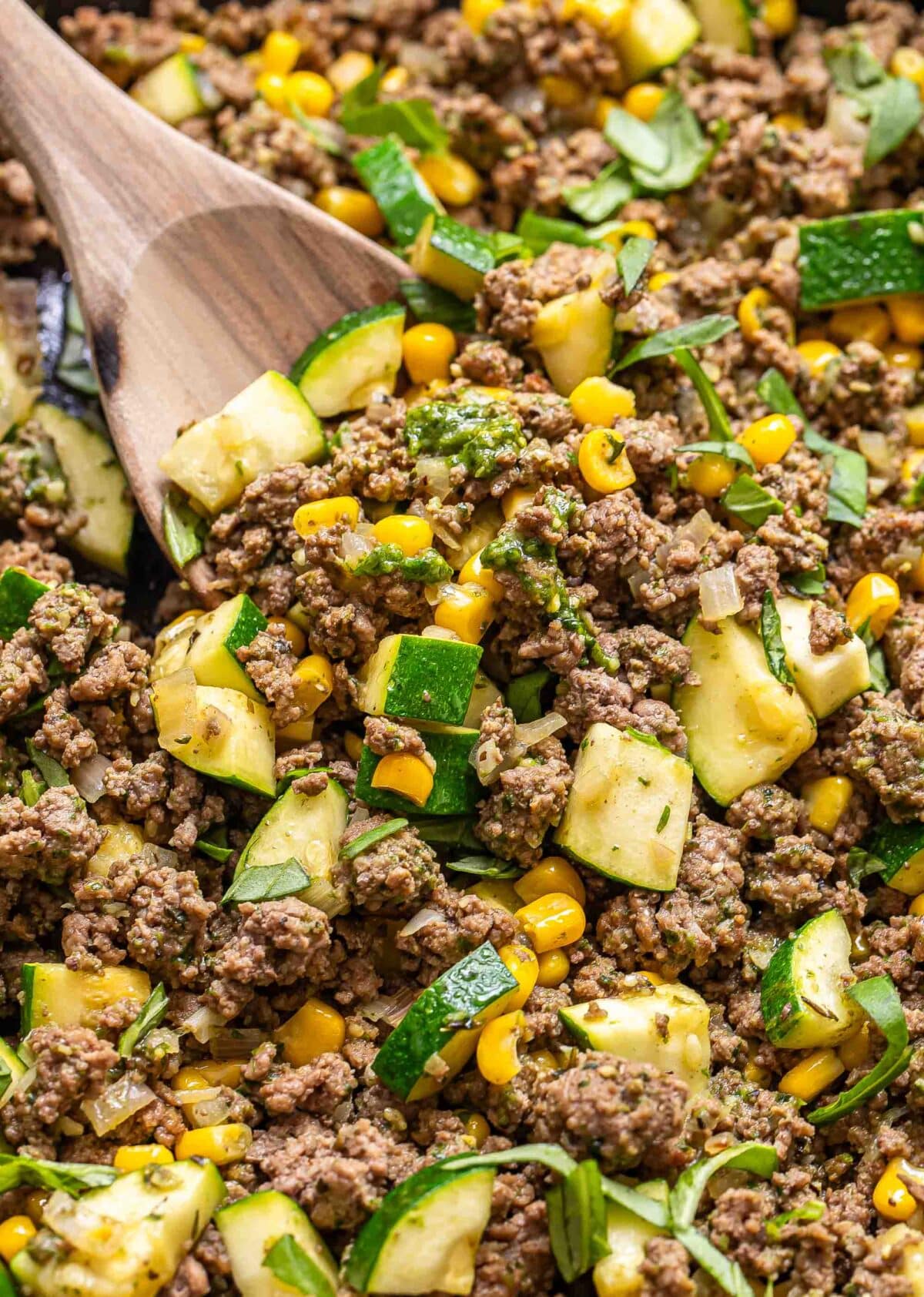Pesto ground beef with zucchini and corn being scooped up with a wooden serving spoon.
