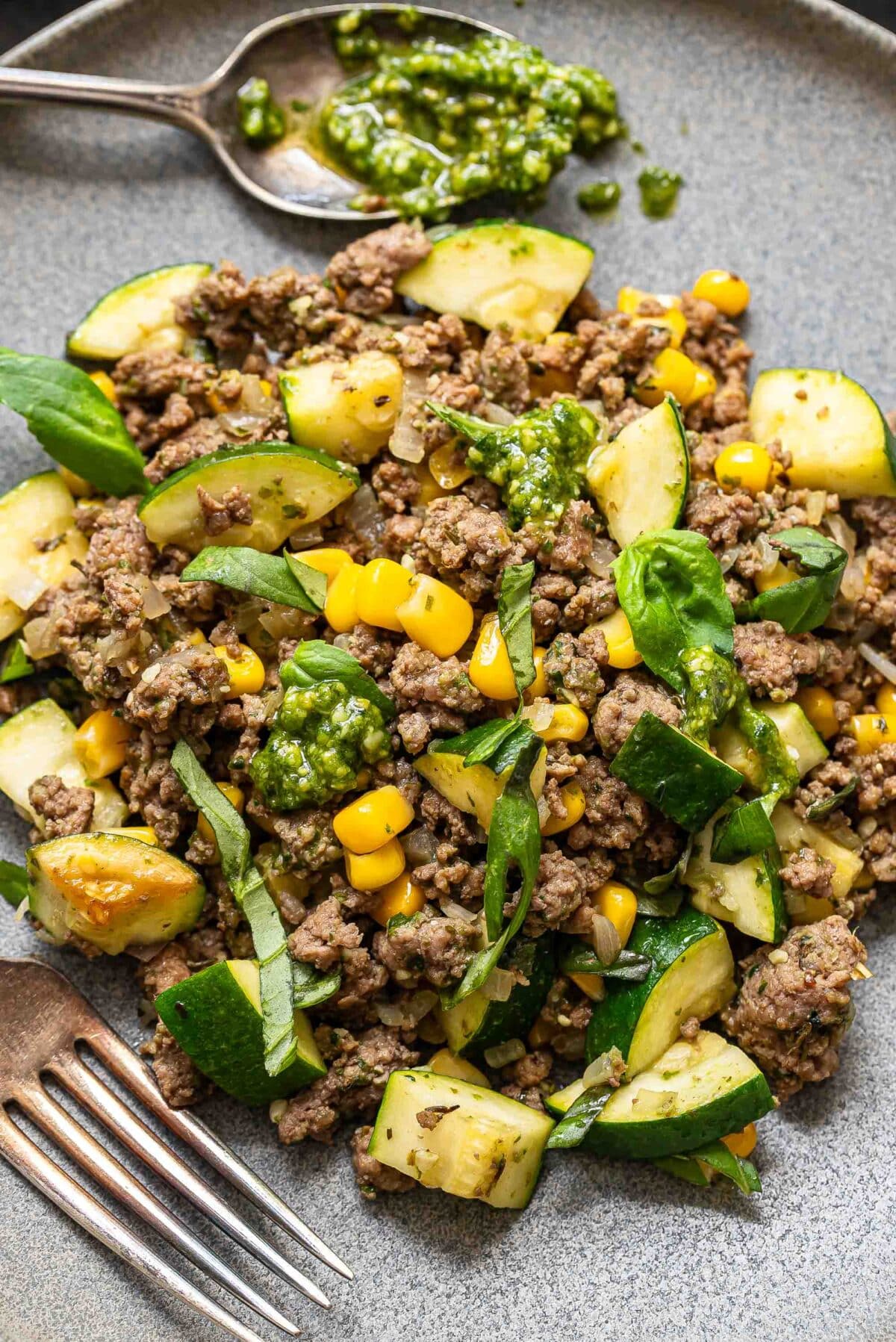 Pesto ground beef with zucchini and corn on a gray plate with a fork and a spoon with pesto on it.