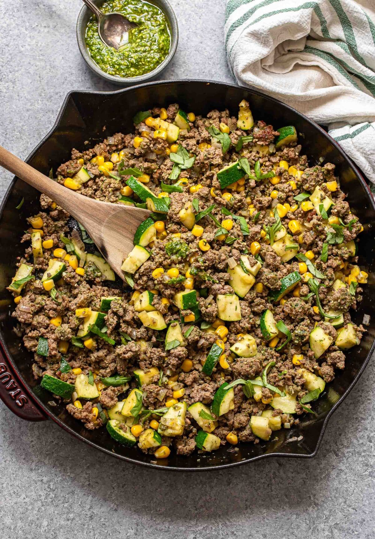 Pesto ground beef with zucchini and corn in a skillet with a wooden serving spoon.
