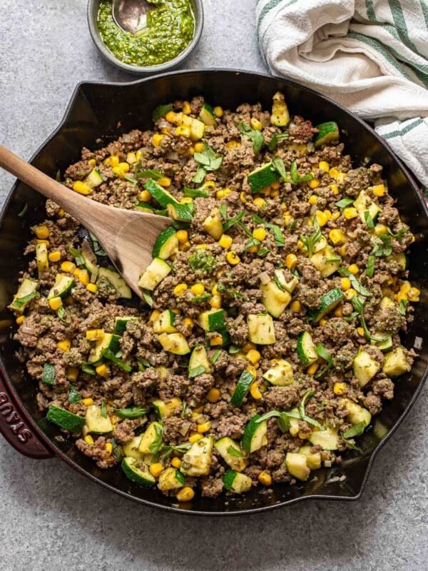 Pesto ground beef with zucchini and corn in a skillet with a wooden serving spoon.
