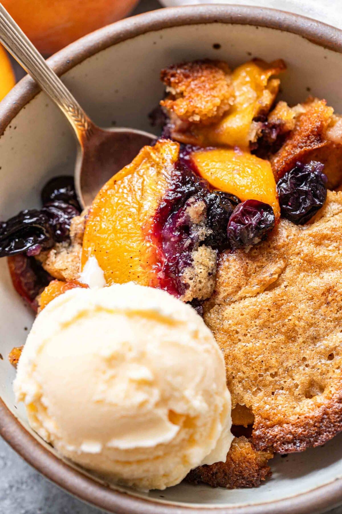 Peach blueberry cobbler in a bowl with a spoon and a scoop of vanilla ice cream.