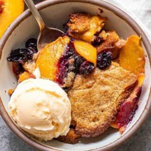 Peach blueberry cobbler in a white bowl with a spoon and a scoop of vanilla ice cream.