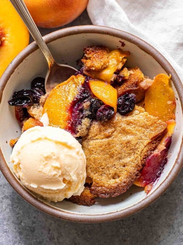 Peach blueberry cobbler in a white bowl with a spoon and a scoop of vanilla ice cream.