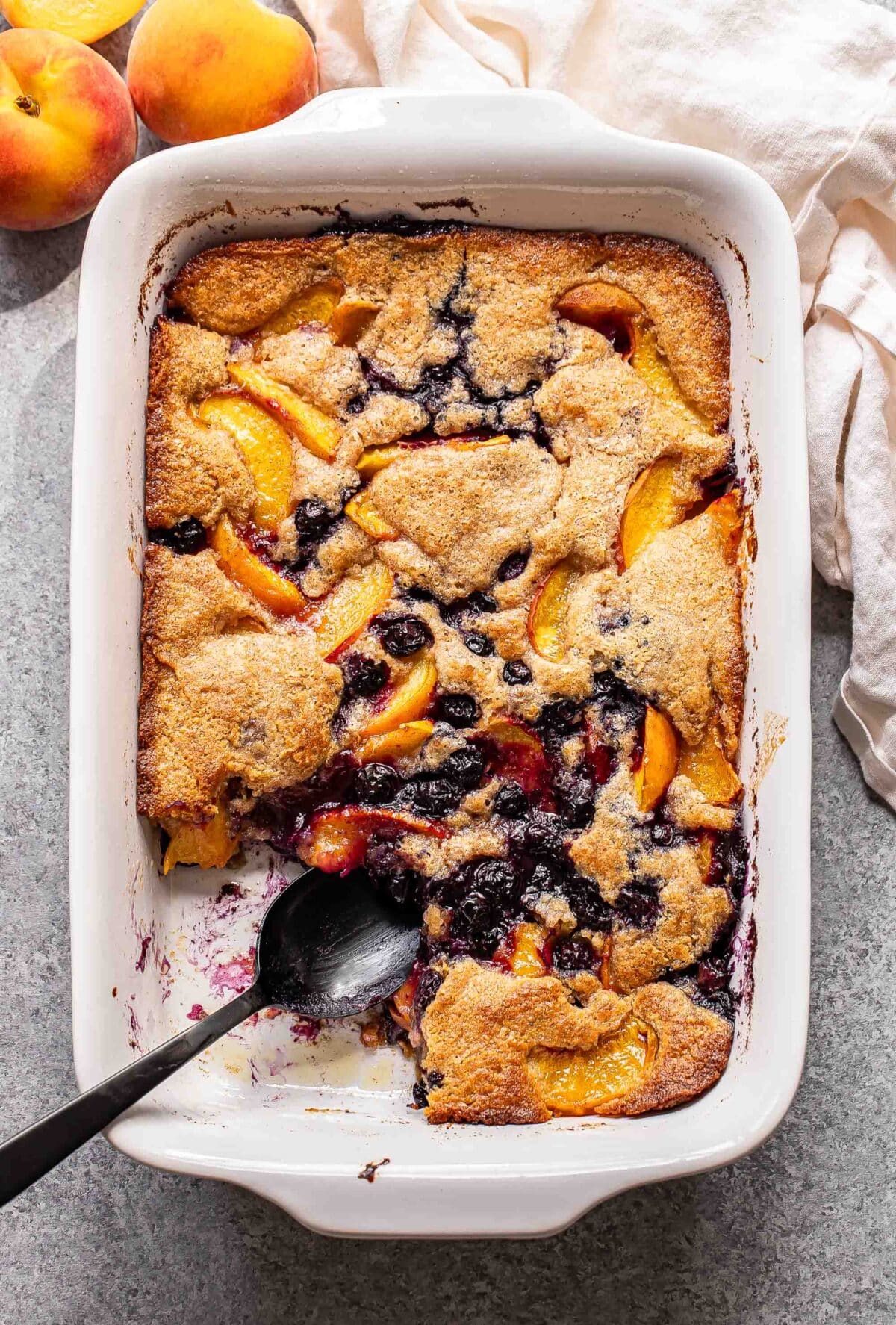 Peach blueberry cobbler in a white rectangular baking dish with a piece missing and a black serving spoon in it's place.