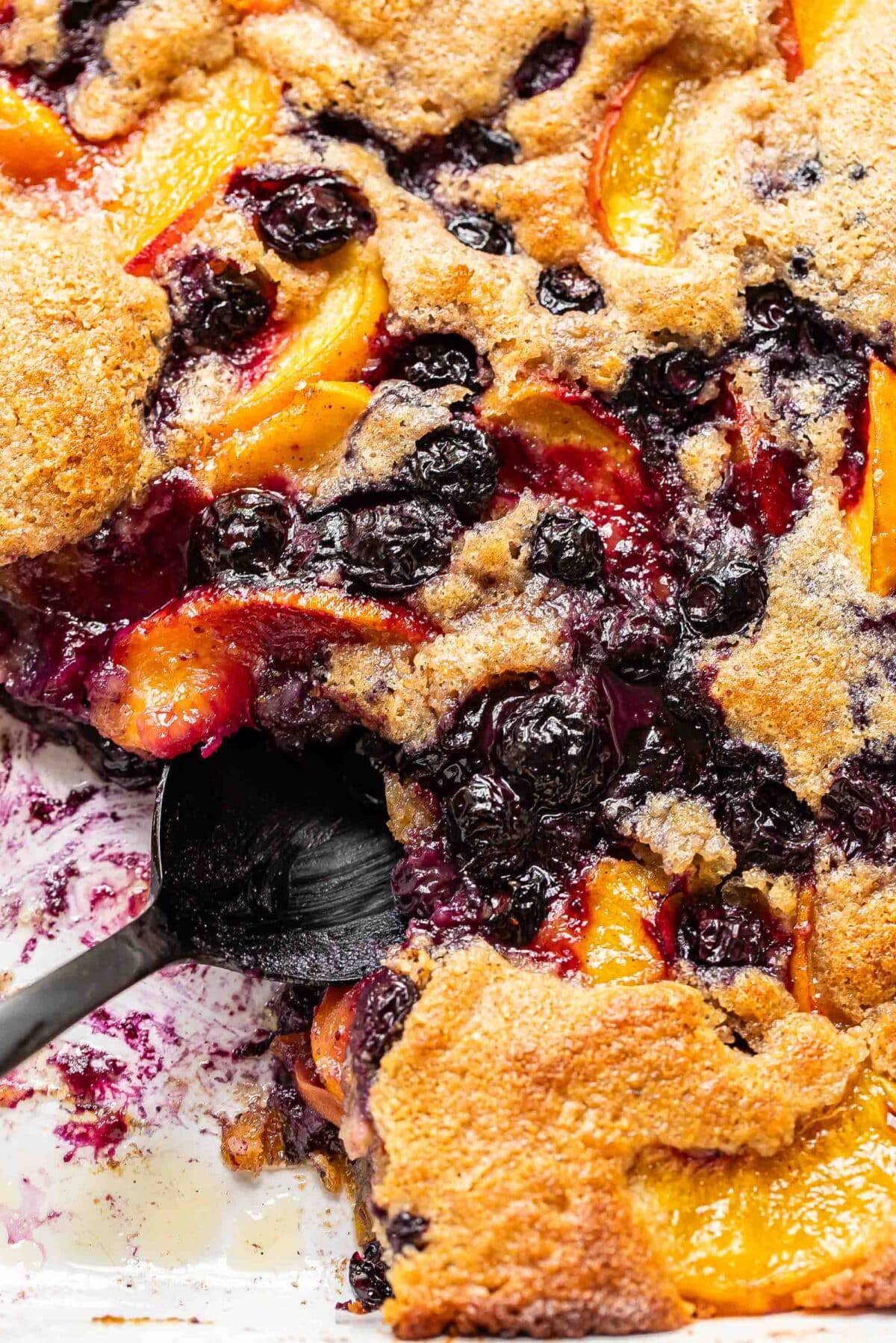Peach blueberry cobbler in a baking dish with a scoop of it cut out and a black serving spoon in it's place.