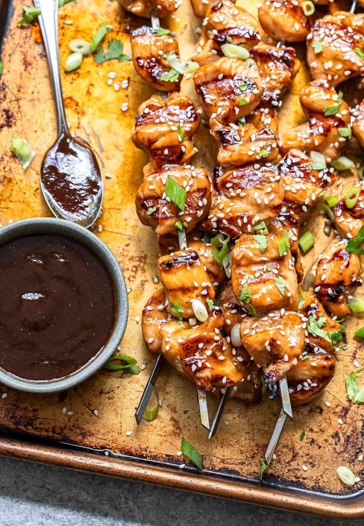 Grilled teriyaki chicken skewers on a sheet pan with a bowl of teriyaki sauce.