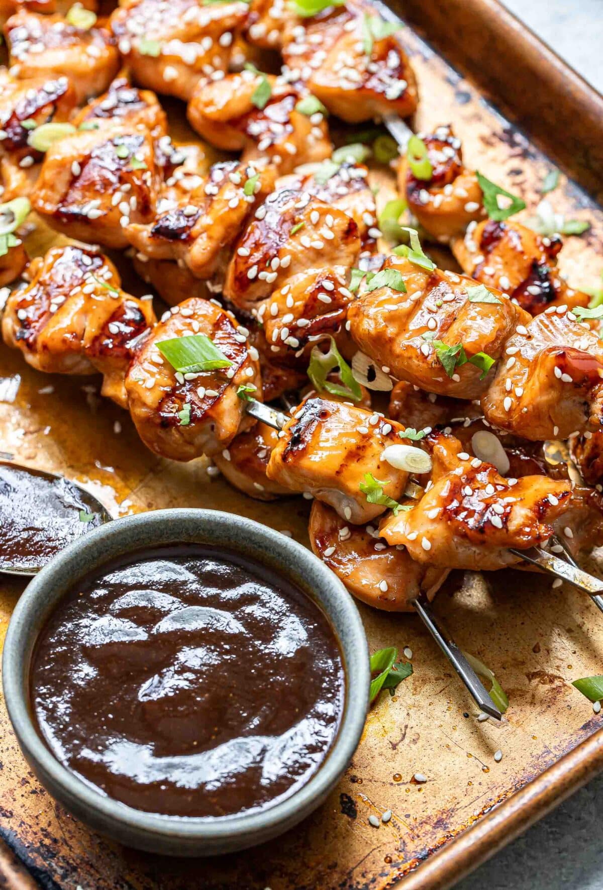 Grilled teriyaki chicken skewers on a sheet pan with a bowl of teriyaki sauce. They're garnished with sesame seeds, green onions, and cilantro.