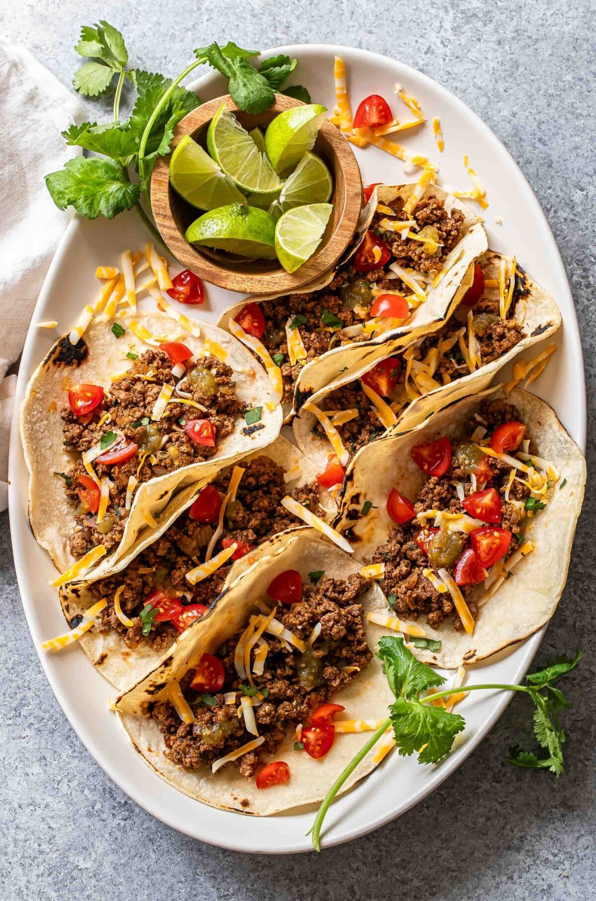6 ground beef tacos on a oval white serving platter with a bowl of lime wedges.