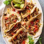 6 ground beef tacos on a oval white serving platter with a bowl of lime wedges.