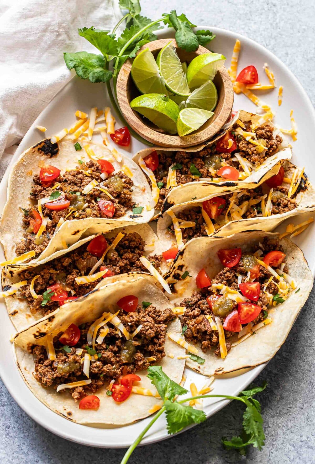 6 ground beef tacos on a oval white serving platter with a bowl of lime wedges.