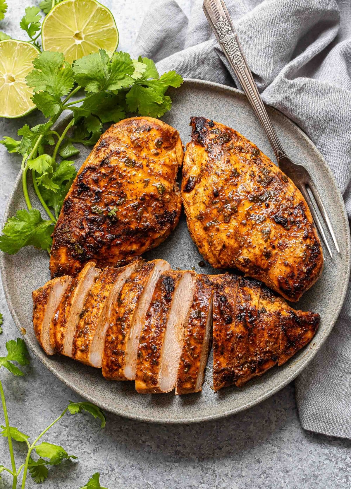 Three southwest grilled chicken breasts on a gray plate with a fork and cilantro. One of the chicken breasts is sliced.