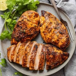 Three southwest grilled chicken breasts on a gray plate with a fork and cilantro. One of the chicken breasts is sliced.