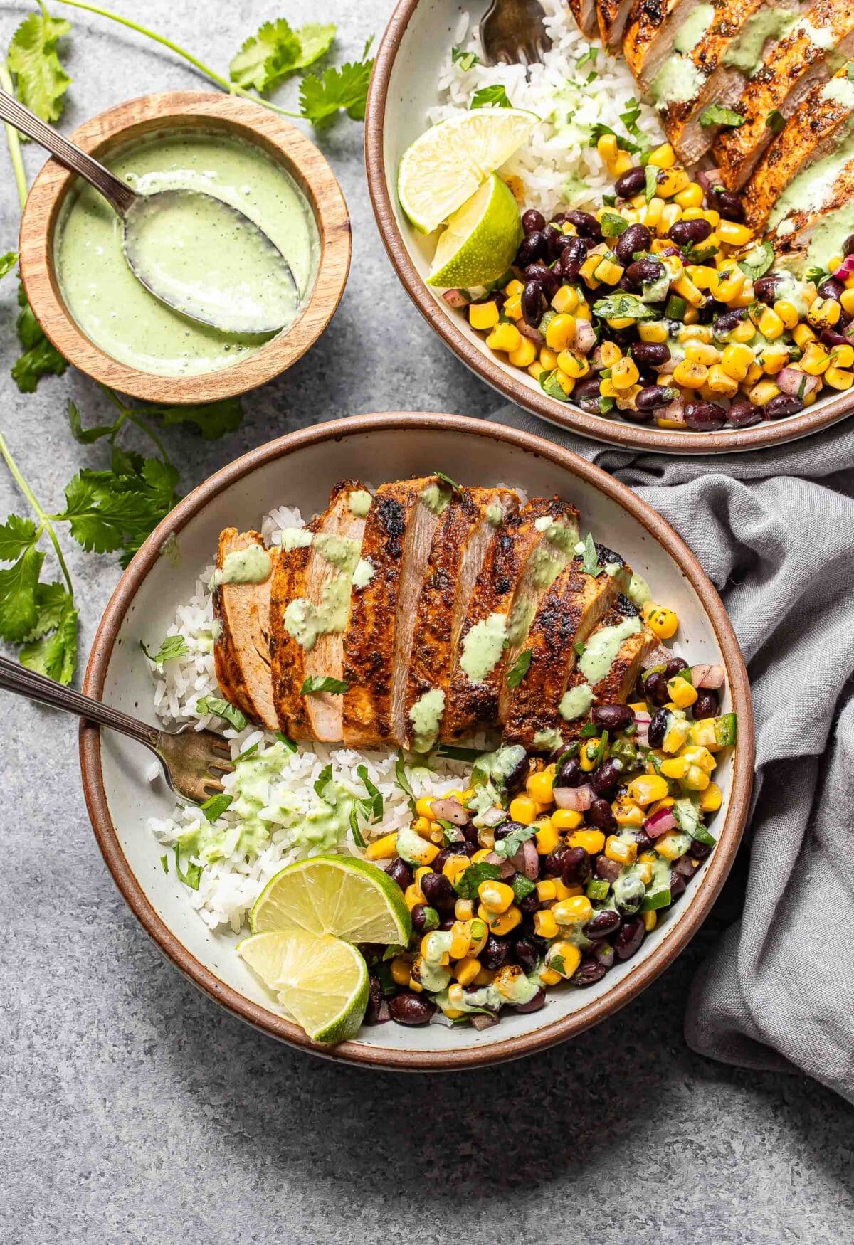 Two Southwest chicken and rice bowls with black bean corn salsa and cilantro lime dressing in a white bowls with forks. A bowl of the dressing is next to it.