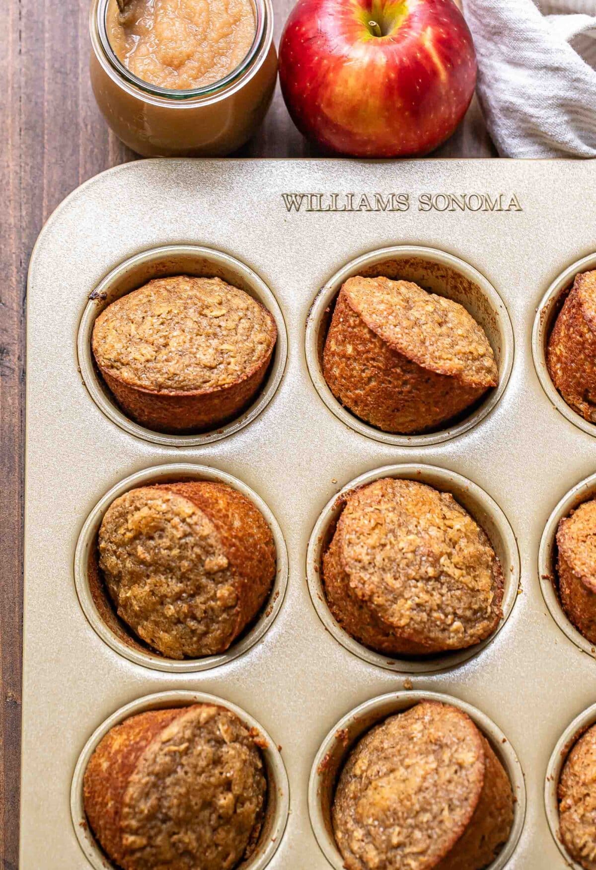 Applesauce muffins in a muffin pan with a jar of applesauce and an apple behind them.