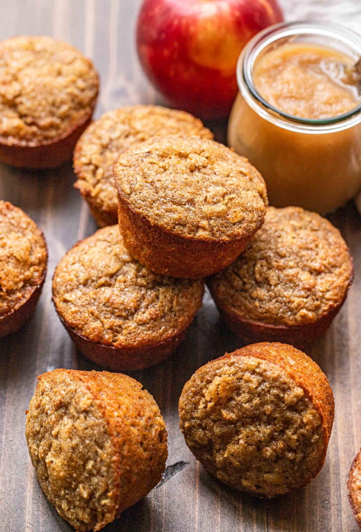 Applesauce muffins stacked on top of each other with a jar of applesauce and an apple behind them.