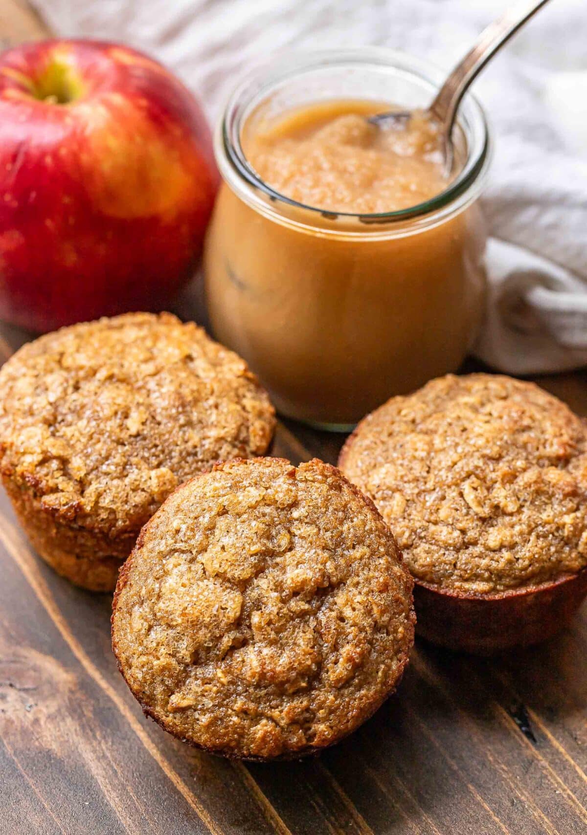 Three applesauce muffins with a jar of applesauce and an apple behind them.