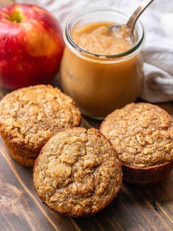 Three applesauce muffins with a jar of applesauce and an apple behind them.
