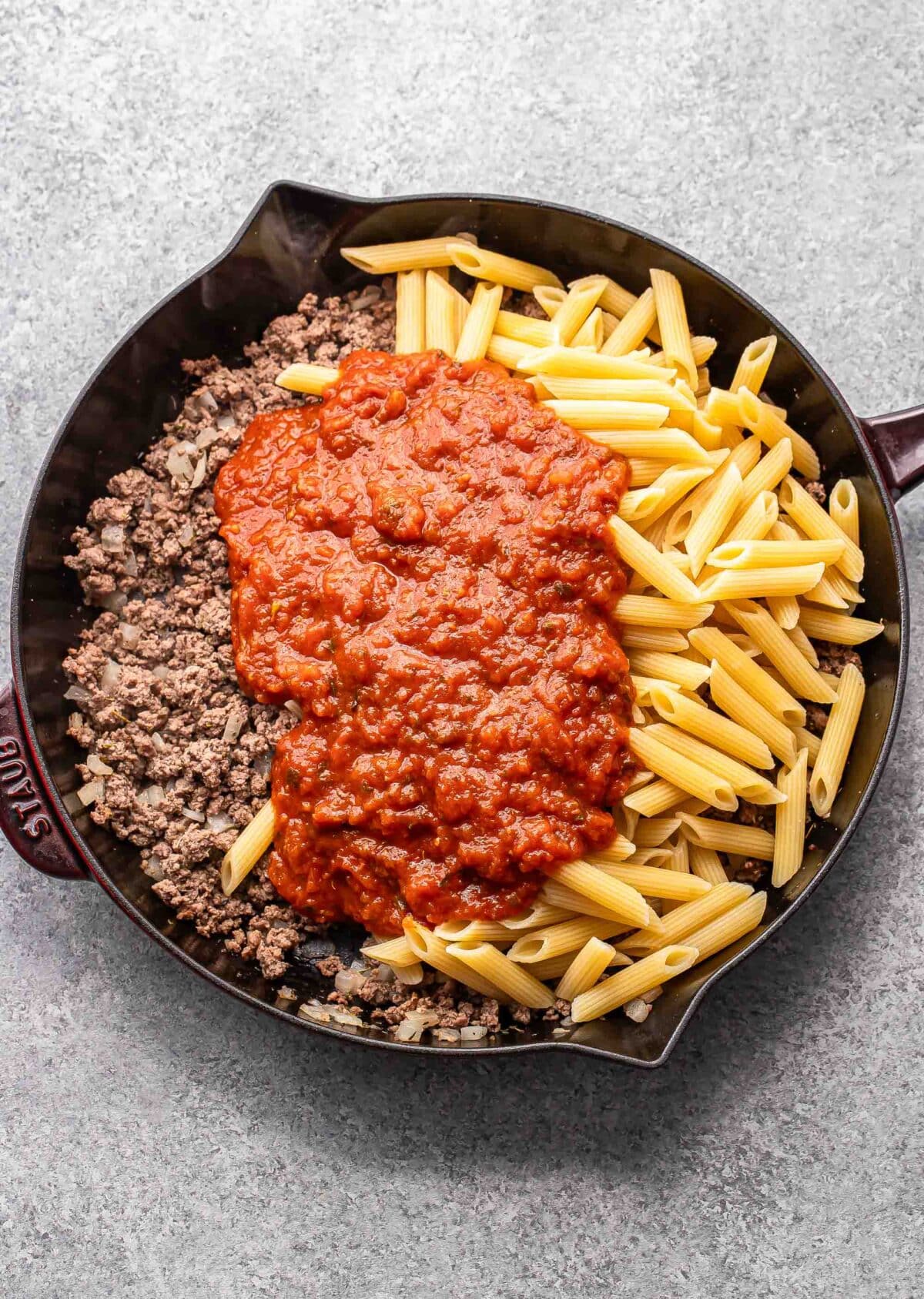 Ground beef, penne, and marinara sauce in a skillet before being stirred together.