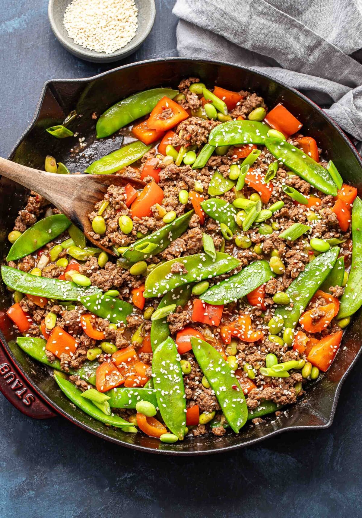 Ground beef stir fry with snow peas, red bell pepper, and edamame in a skillet with a wooden serving spoon.
